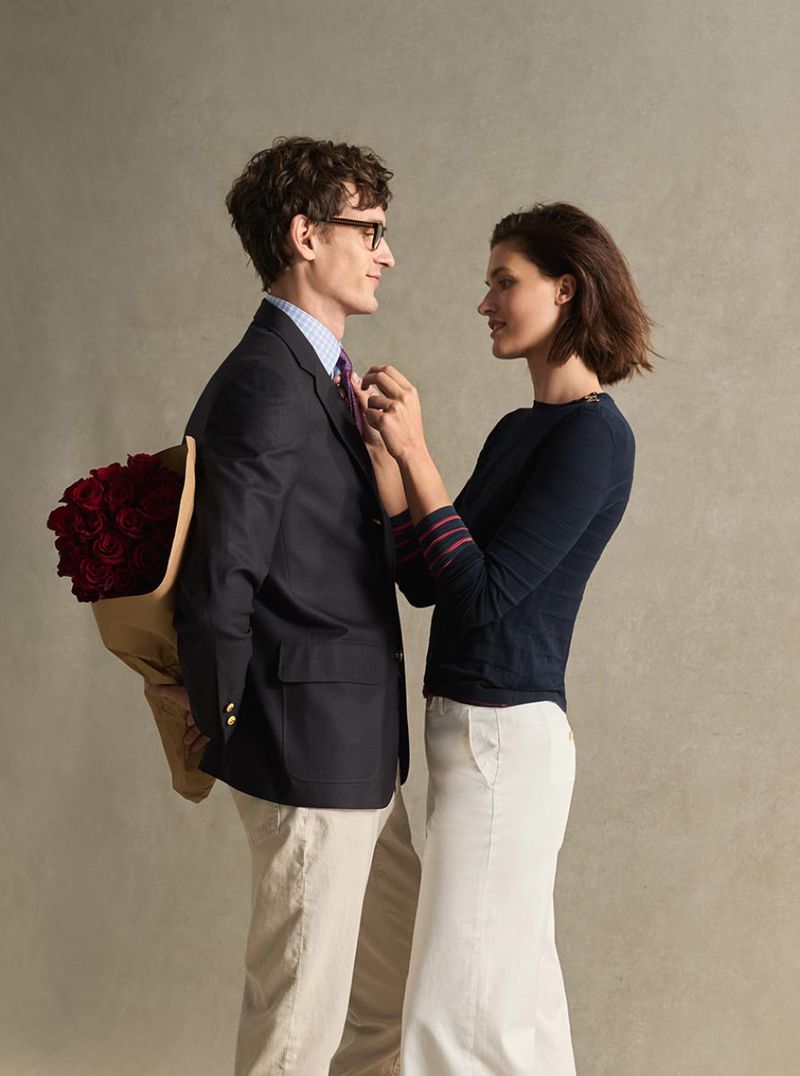 Man in navy blazer and woman in dark sweater facing each other, holding a bouquet of red roses behind his back