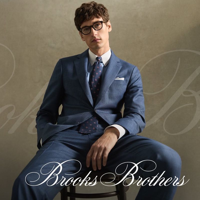 Model wearing a blue suit with patterned tie and glasses seated against a textured wall for Brooks Brothers