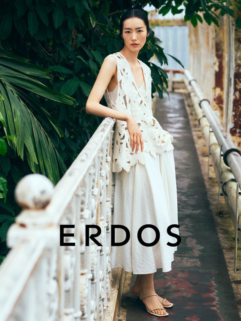 Model Liu Wen leaning on a white metal railing wearing a white sleeveless cutout top and long skirt in a lush garden