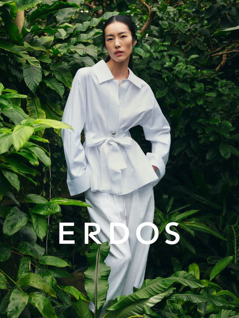 Model Liu Wen wearing a white buttoned shirt and matching trousers standing amidst lush tropical foliage for Erdos Spring Summer 2026.