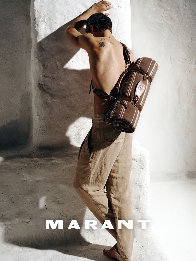 Shirtless male model wearing neutral trousers leaning against a white textured wall carrying a rolled blanket strap bag