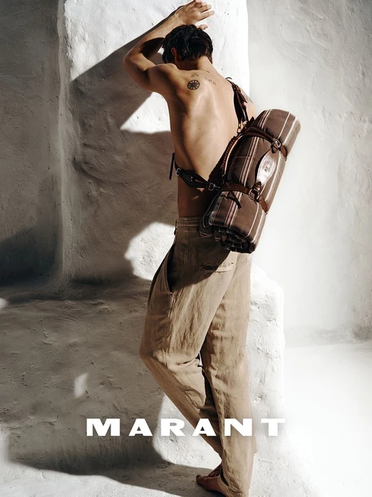Shirtless male model wearing neutral trousers leaning against a white textured wall carrying a rolled blanket strap bag