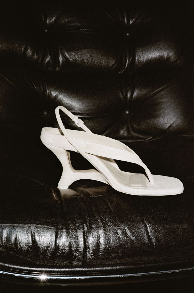 White slingback sandal with sculpted heel resting on a black tufted leather armchair for Simkhai S/S 26 lookbook