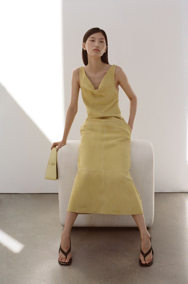 Model seated on a white block wearing a yellow top and matching midi skirt holding a small handbag