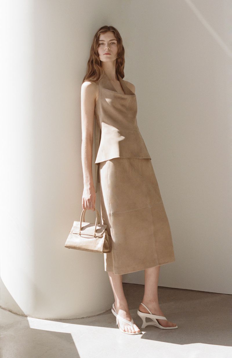 A slender woman models tan halter top and skirt with a tan purse and white heels against a neutral wall and sunlight in studio.