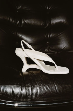 White slingback sandal with sculpted heel resting on a black tufted leather armchair for Simkhai S/S 26 lookbook