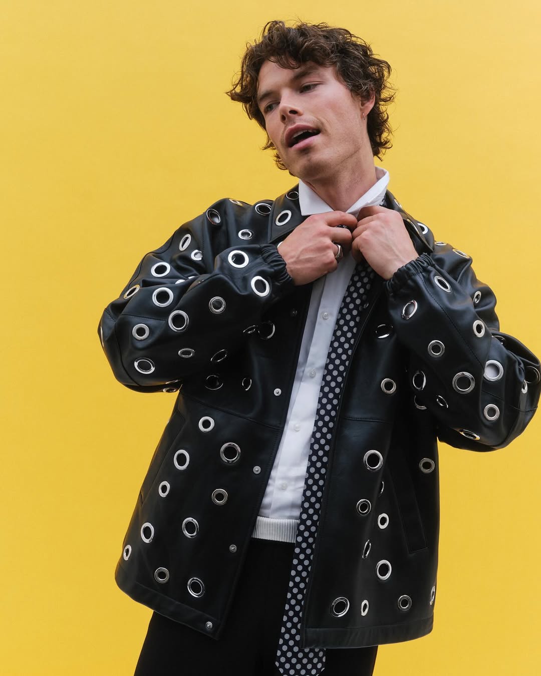 Young man in a black grommeted jacket adjusts his collar against a vibrant yellow backdrop, showcasing a modern fashion statement.