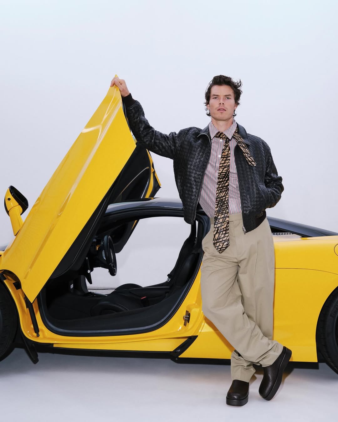 Stylish man in a jacket, tie, and khaki pants poses next to a bright yellow sports car with an open door against a plain background.