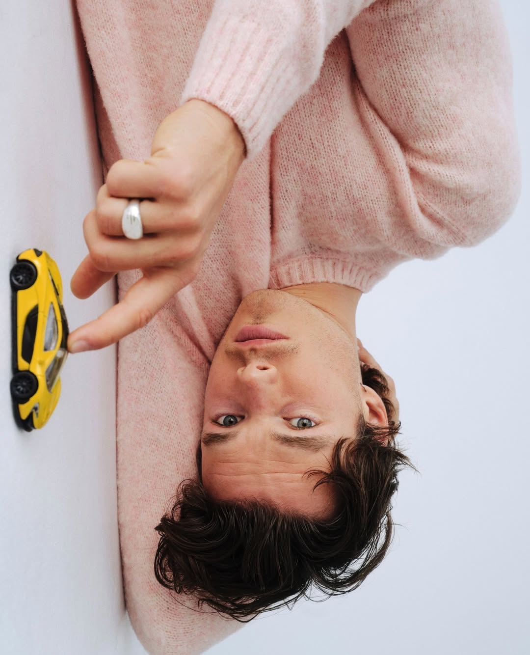 Adult male in a pink sweater poses with a yellow toy car against a white background in a studio with vertical composition.