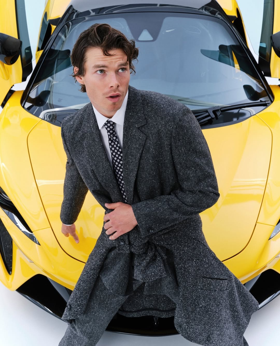 Adult male in a grey overcoat poses in front of a bright yellow sports car with doors open, creating a high-fashion automotive scene.