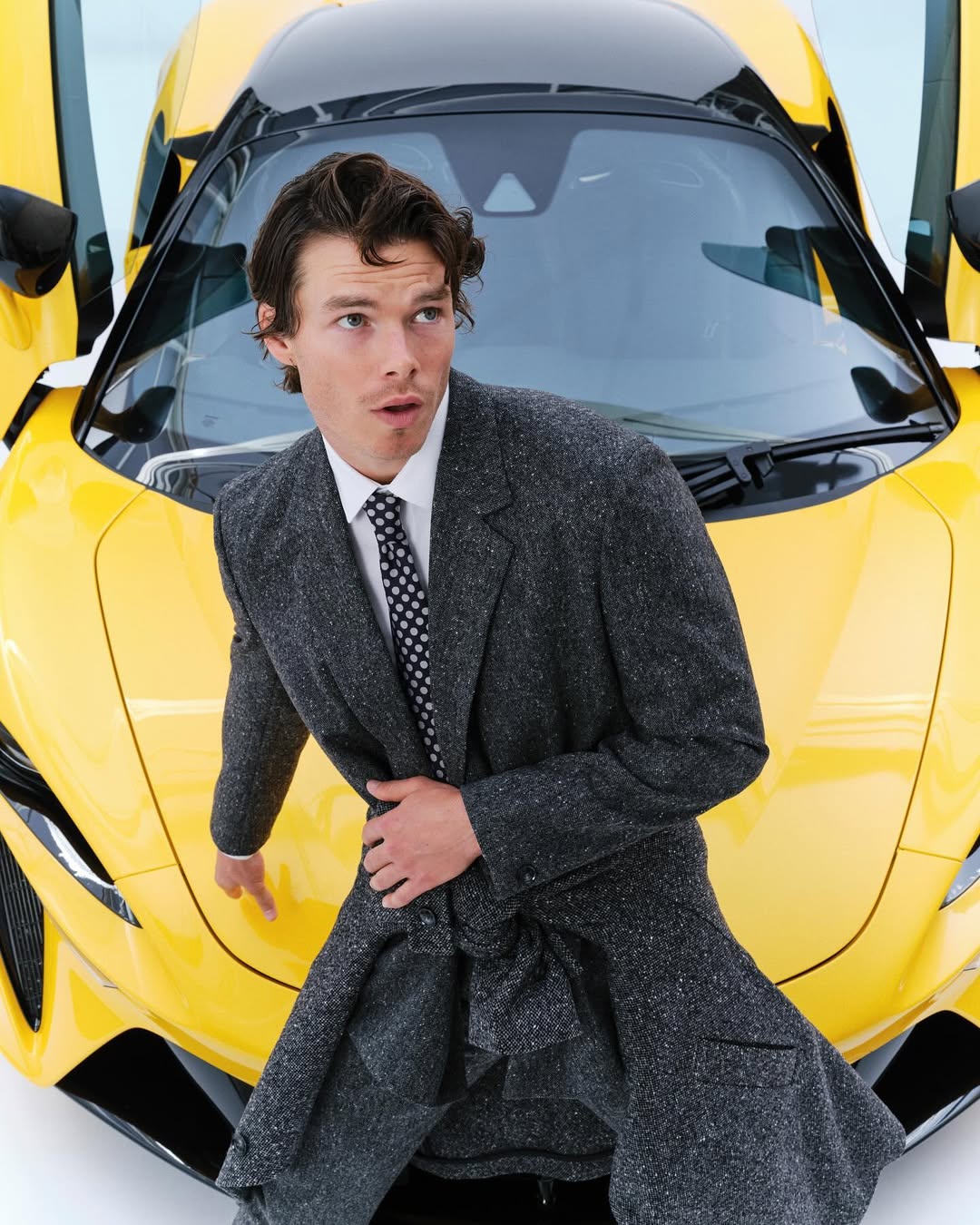 A stylish man in a gray coat stands beside a bright yellow sports car with its door open, against a clean white backdrop.