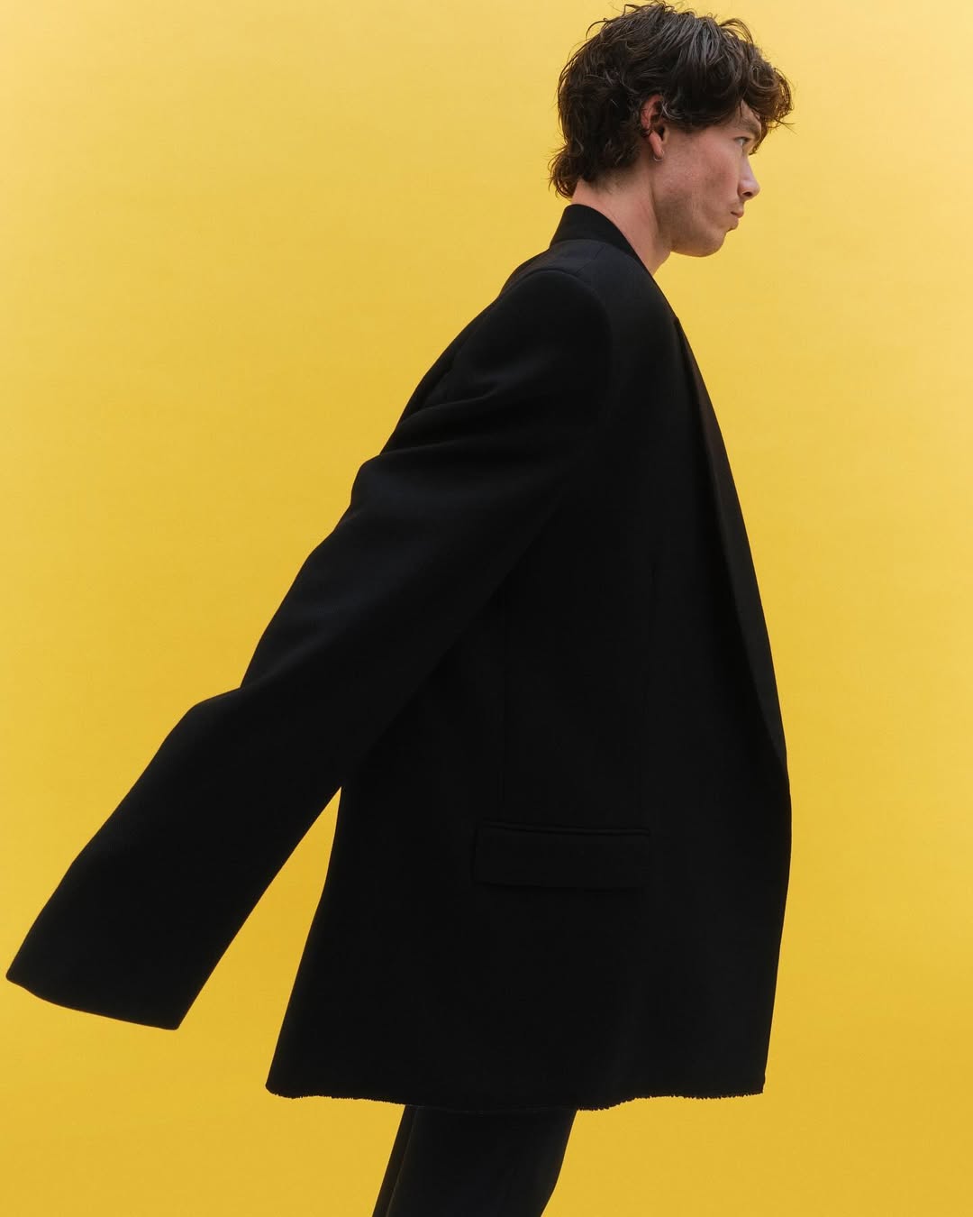 Profile shot of a slender adult male in a dark suit against a solid yellow background, showcasing minimalist fashion and studio lighting.