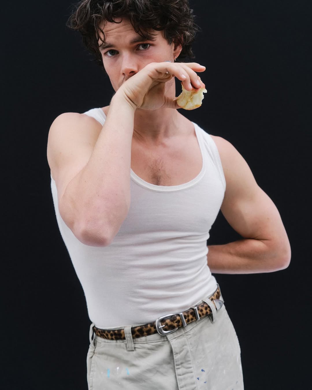 Sam Corlett wearing a white tank top and patterned belt holding a bitten piece of fruit against a black seamless studio background.