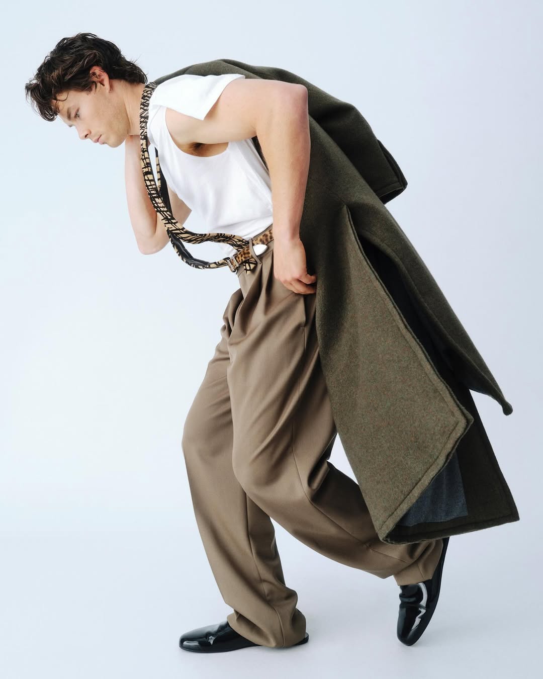 Fashionable man wearing a white t-shirt, brown dress pants, and black shoes, carrying a green wool coat over his shoulder in a studio.