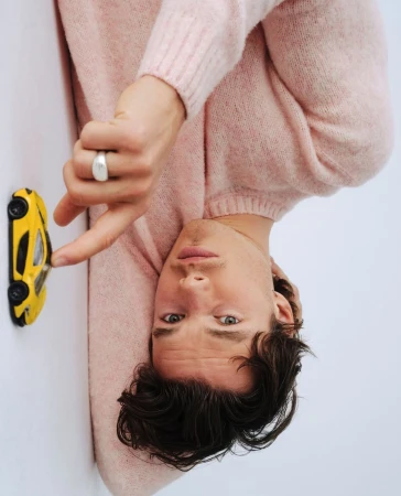 Close-up portrait of a man wearing a pink knit sweater pointing at a small yellow toy sports car against a white studio background.