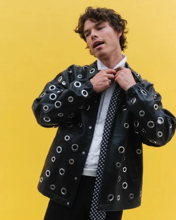 Sam Corlett wearing a black jacket with metal grommets and a patterned tie against a solid yellow background