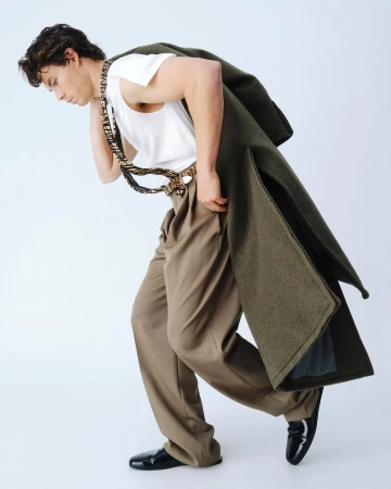 Sam Corlett leaning forward in a white tank top and brown trousers with an olive coat draped over his shoulders against a white background.