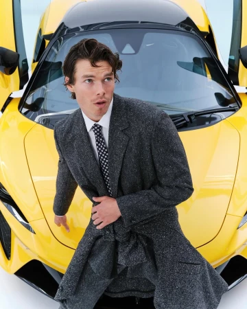 Sam Corlett wearing a grey tweed coat over a white shirt and polka dot tie in front of a yellow sports car
