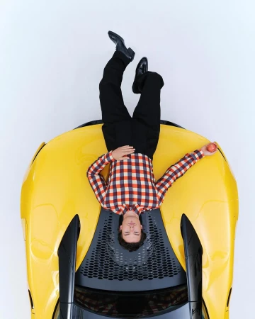 Man wearing a plaid shirt and dark trousers lying on the hood of a bright yellow sports car against a white background