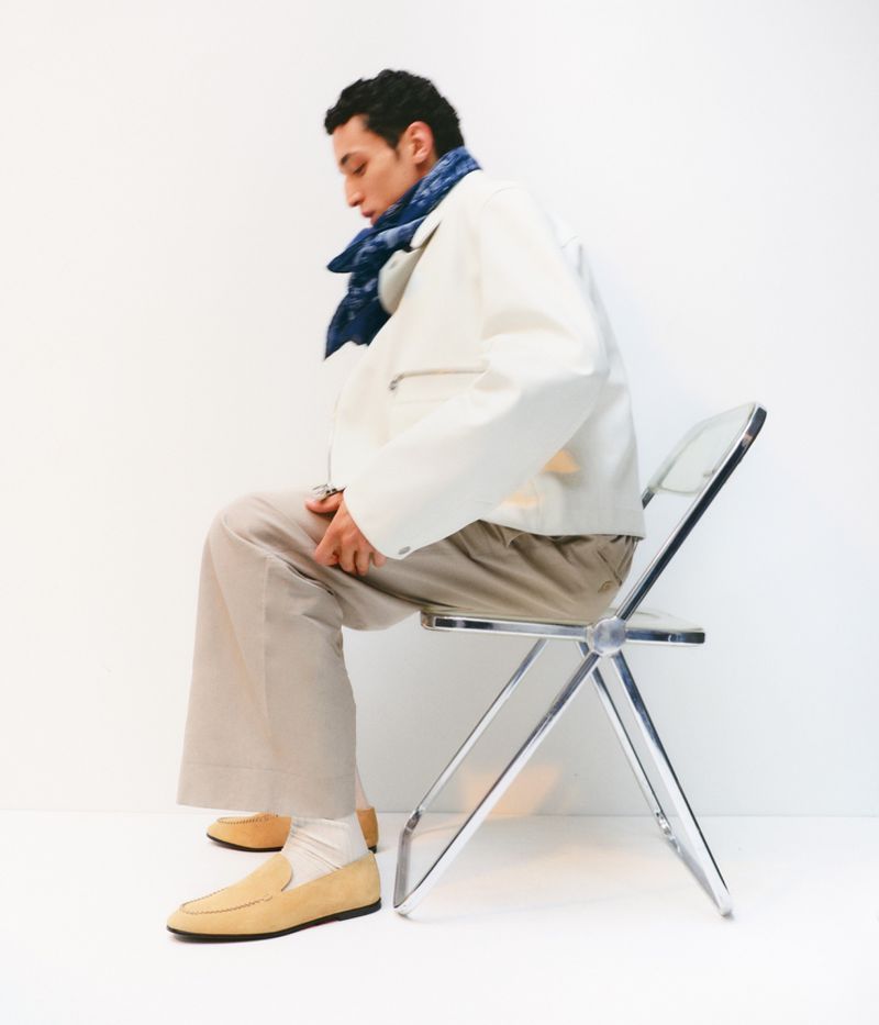 An adult male model sits in a clear chair wearing a white jacket, beige trousers, tan suede loafers, and a blue patterned scarf. 