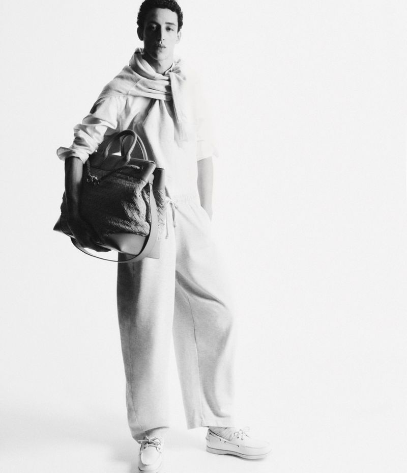 Black and white studio shot of a young adult male model wearing loose-fitting casual clothing, holding a tote bag, and posing.