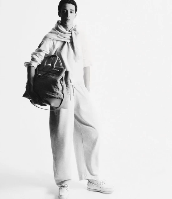 Black and white studio shot of a young adult male model wearing loose-fitting casual clothing, holding a tote bag, and posing.