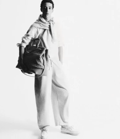 Black and white studio shot of a young adult male model wearing loose-fitting casual clothing, holding a tote bag, and posing.
