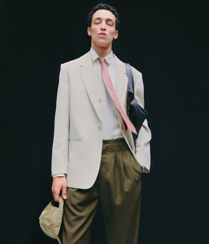 Adult male in light beige suit with pink tie against dark backdrop. He holds a tan cap and carries a black bag over his shoulder.
