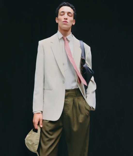 Adult male in light beige suit with pink tie against dark backdrop. He holds a tan cap and carries a black bag over his shoulder.