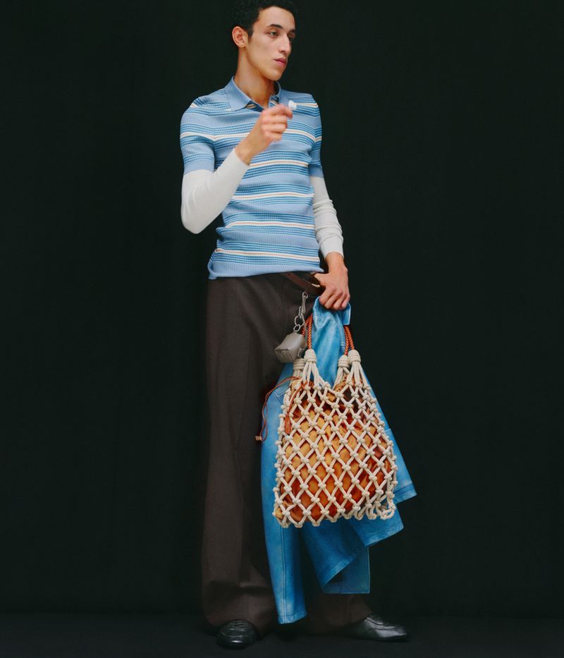 A slender adult male model in a blue striped polo shirt and brown wide-leg trousers holds a white net bag in a full shot against a black background.