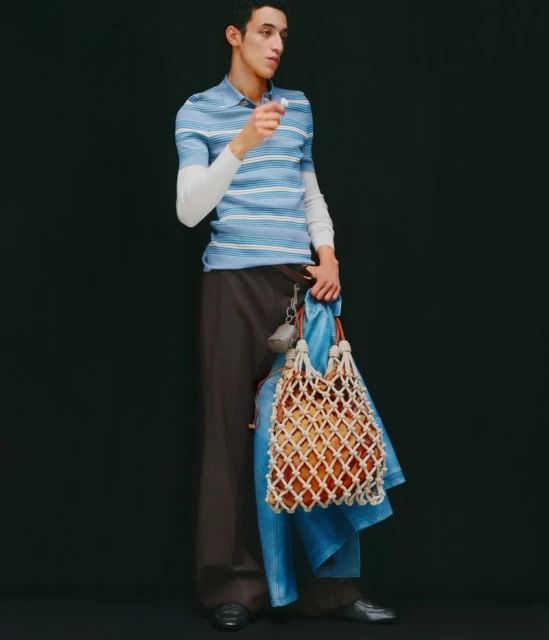 A slender adult male model in a blue striped polo shirt and brown wide-leg trousers holds a white net bag in a full shot against a black background.