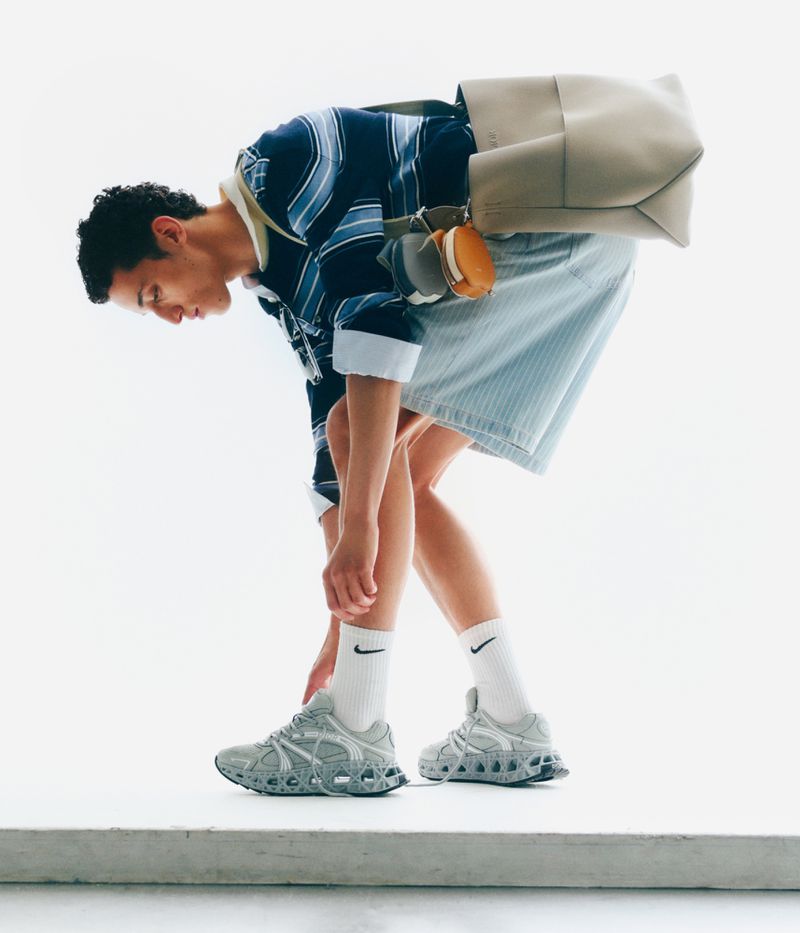 Man in athletic wear bends to tie his shoelaces indoors, wearing a striped rugby shirt, shorts, socks, and grey running shoes, and carrying a bag.