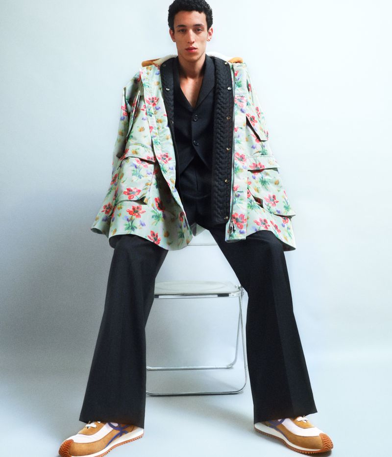 Young adult posing on a chair in a floral jacket, black vest and pants, and athletic shoes, set against a simple gradient background.