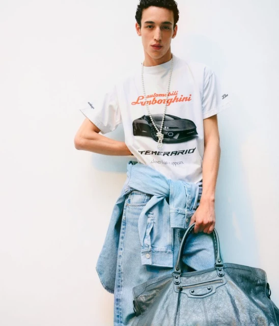 Man in Lamborghini T-shirt with jeans tied around his waist, holding a large bag against a white wall, studio lighting.