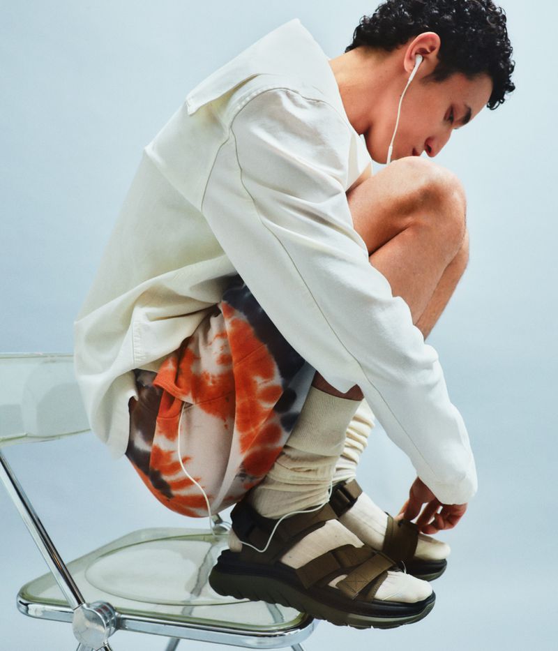 A male model crouches on a clear chair, wearing sandals, patterned shorts, a cream jacket, and earbuds against a light blue background.