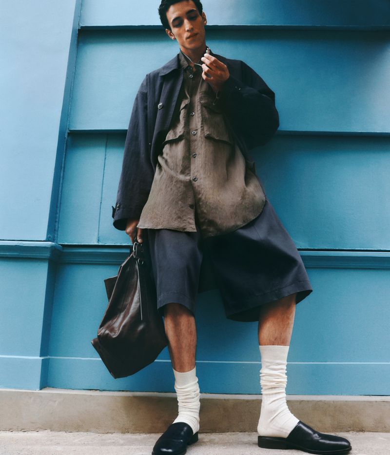 Man in casual fashion posing against a blue wall. He wears shorts, a shirt, and a jacket, holding a bag with loafer shoes and white socks.