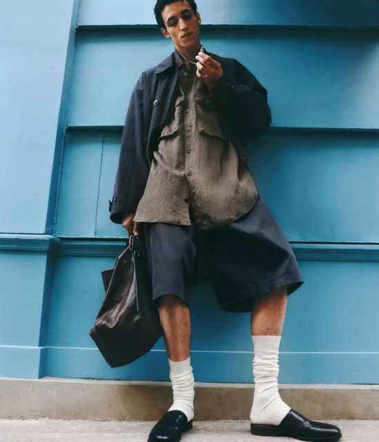 Man in casual fashion posing against a blue wall. He wears shorts, a shirt, and a jacket, holding a bag with loafer shoes and white socks.