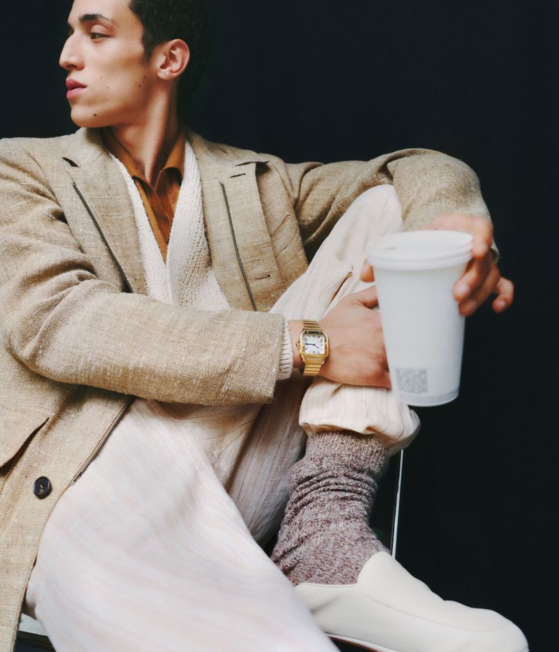 Man in a beige suit holds a white cup, wearing a gold watch and white loafers, posing against a dark background in a studio shot.