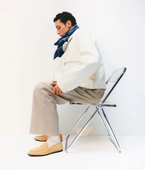 Man seated on a chrome folding chair wearing a white jacket, beige trousers, loafers, and a blue patterned scarf against a plain white background