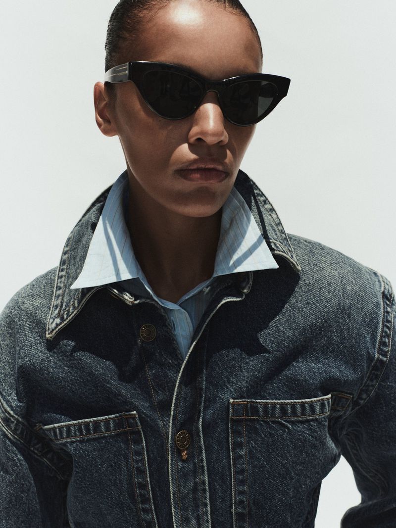 Model wearing dark sunglasses, denim jacket and light blue shirt against a white background for AGOLDE Spring 2026