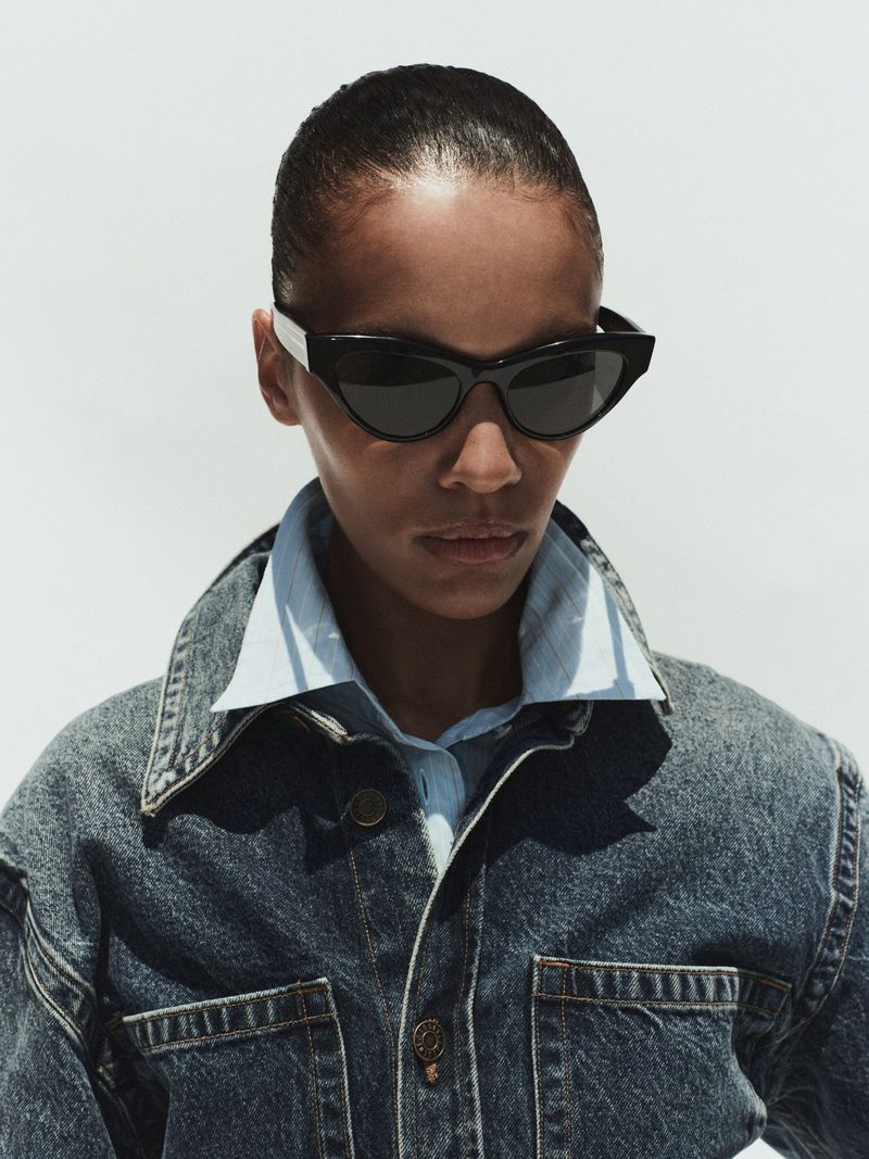 Model wearing dark sunglasses, a light blue collared shirt, and a denim jacket against a bright white background.