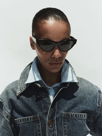 Model wearing dark sunglasses, a light blue collared shirt, and a denim jacket against a bright white background.