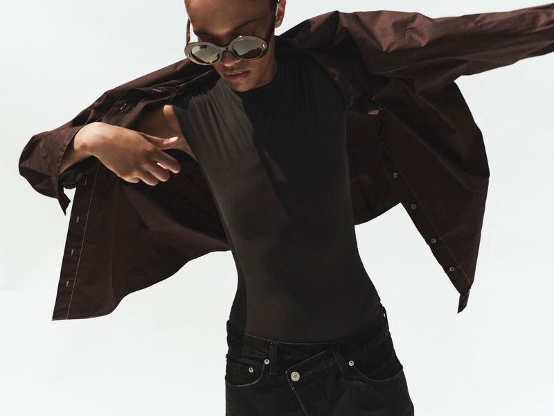 Model wearing dark sunglasses and an open brown shirt over a black top against a plain white background.