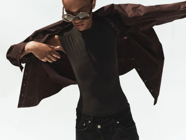 Model wearing dark sunglasses and an open brown shirt over a black top against a plain white background.
