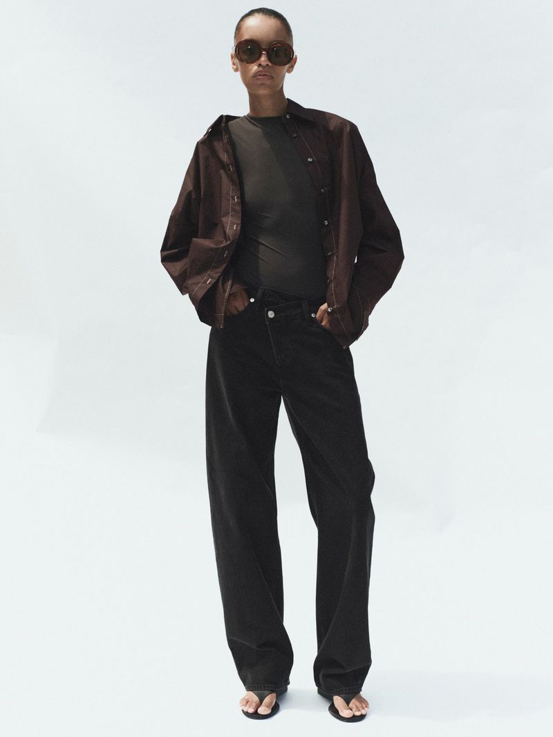 Model wearing dark sunglasses, brown button-down shirt, and black jeans against a white background.
