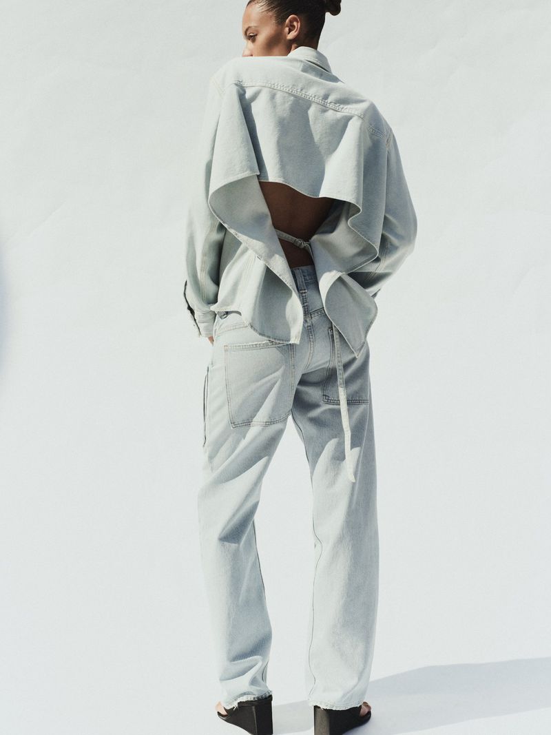Back view of a person wearing a light blue denim jacket with an open back, matching jeans, and black platform shoes against white.