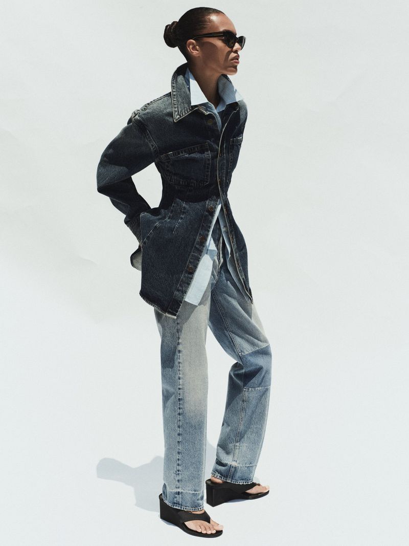 Model wearing a dark denim jacket and light wash jeans standing against a plain white background for AGOLDE.