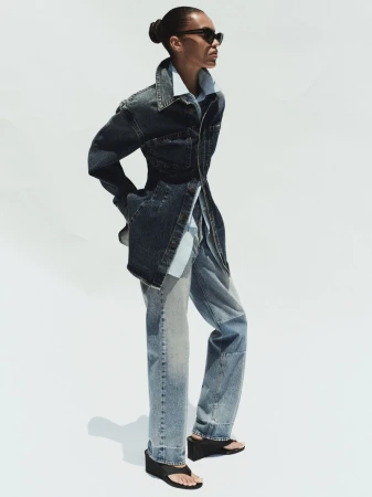 Model wearing a dark denim jacket and light wash jeans standing against a plain white background for AGOLDE.