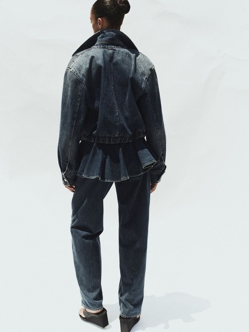Model wearing dark denim jacket and matching jeans seen from behind against a plain white studio background.