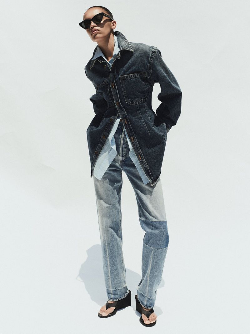 Model in dark sunglasses wearing a denim jacket and jeans against a white seamless background